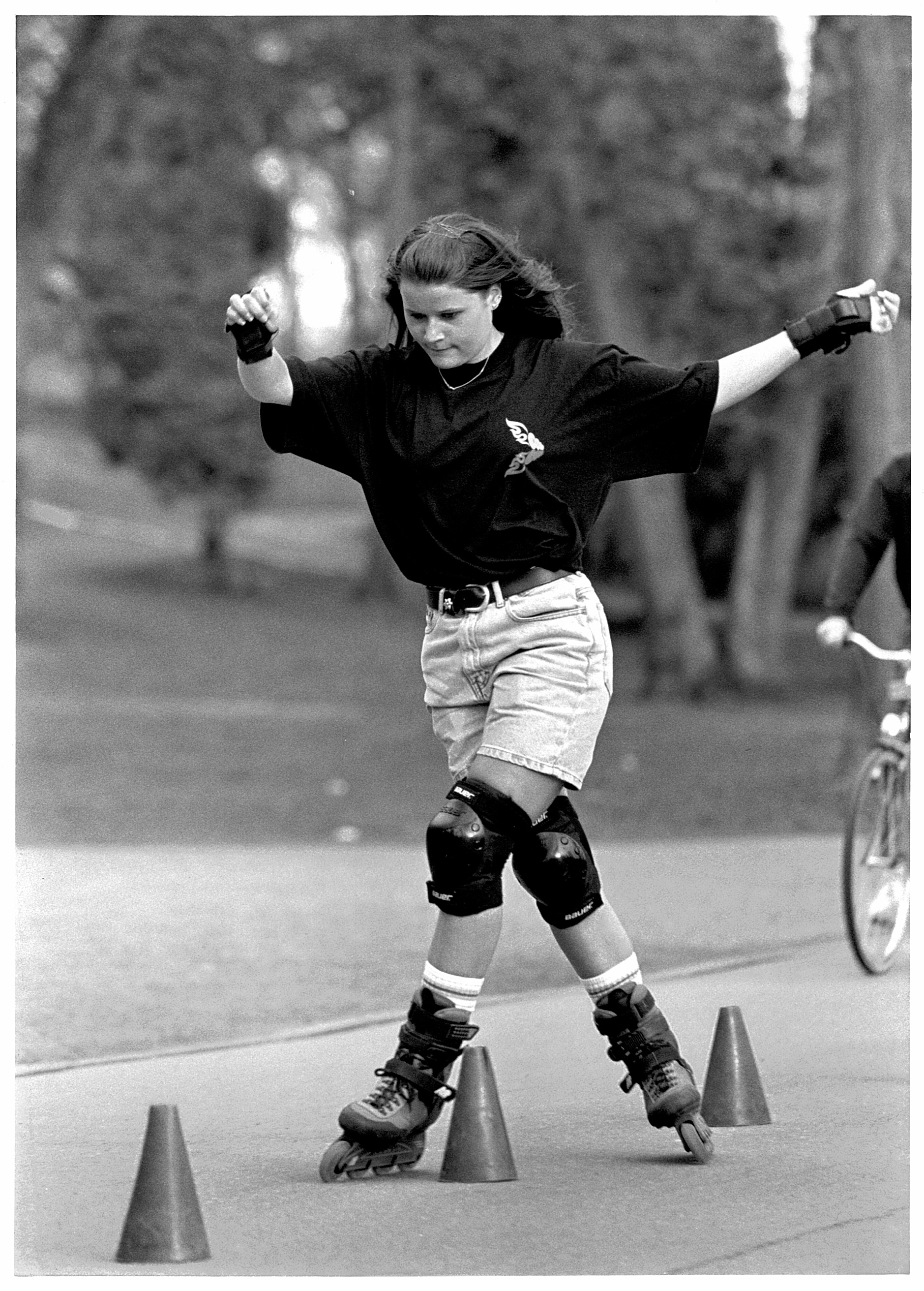 Rullaluistelija. Nuori nainen rullaluistelee ulkona pujotellen muovikartioiden välitse. Hänellä on yllään musta paita ja vaaleat farkkusortsit, joissa musta vyö. Varustukseen kuuluvat myös polvi- ja rannesuojat. Nuorisotyö-lehden kuva. Kuvaaja: Antero Aaltonen. FINNA. 